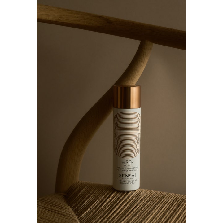 Sensai Silky Bronze Cooling Protective Suncare Spray 150 ml viilentävä aurinkosuoja SPF50 valkoisessa pullossa puisella tuolilla.