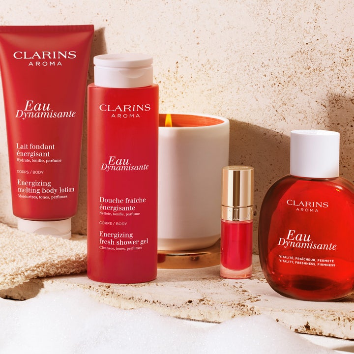 Clarins Eau Dynamisante Energizing Fresh Shower Gel 200 ml suihkugeeli ja muita punaisia Clarins-tuotteita kynttilän rinnalla.