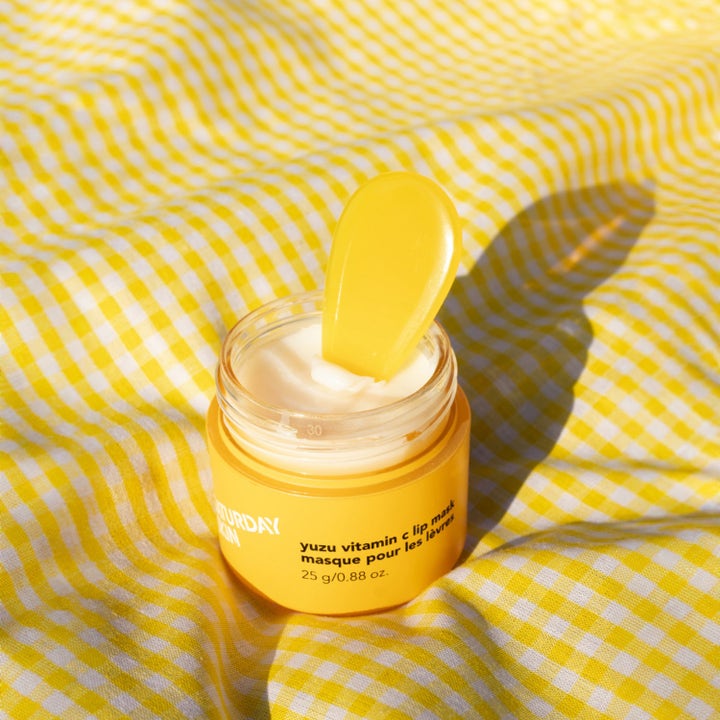Saturday Skin Yuzu Vitamin C Lip Mask 25 g kirkastava huulinaamio keltaisessa purkissa keltaisella ruutukankaalla.