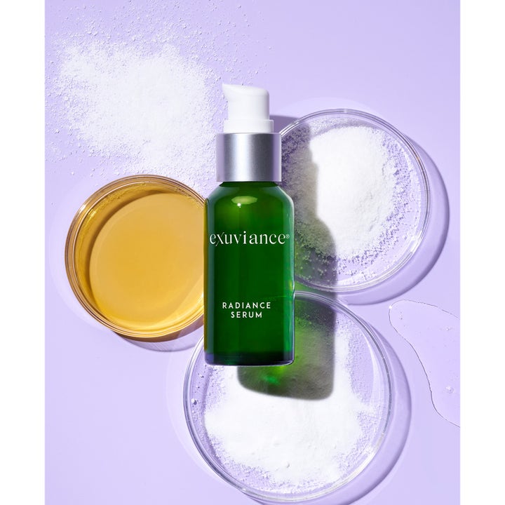 Exuviance Radiance Serum 30 ml heleyttävä seerumi vihreässä lasipullossa petri-maljojen ja ainesosien keskellä.