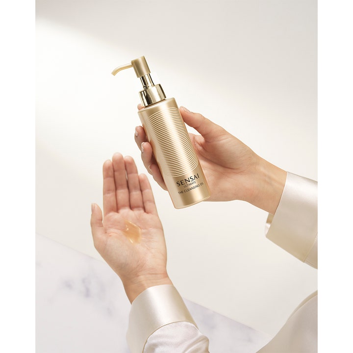 Sensai Ultimate The Cleansing Oil 150 ml puhdistusöljy kultaisessa pullossa ja kämmenellä näkyvä täyteläinen öljy.