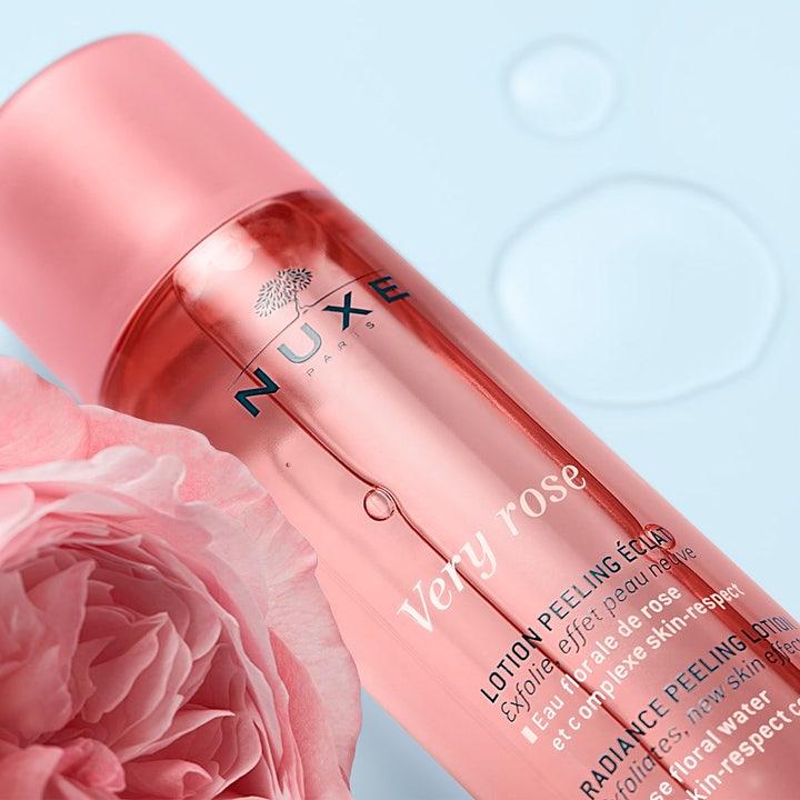 Nuxe Very Rose Peeling Lotion 150 ml kirkastava kuorinta vaaleanpunaisessa pullossa ruusun rinnalla.