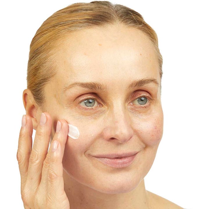 No7 Lift & Luminate Triple Action Night Cream 50 ml yövoide jota nainen levittää poskelleen kirkastamaan ihoa.