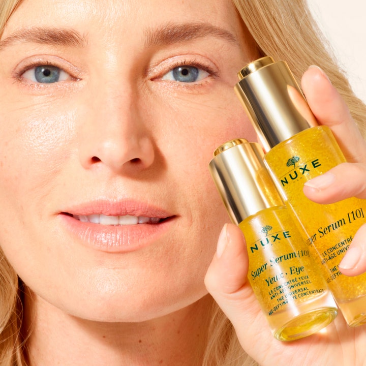 Nuxe Super Serum [10] Eye 15 ml silmänympärysseerumi ja nainen pitelemässä kultaisia seerumipulloja.