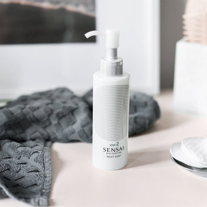 Sensai Silky Purifying Milky Soap kosteuttava puhdistustuote valkoisessa pumppupullossa harmaan pyyhkeen vieressä.