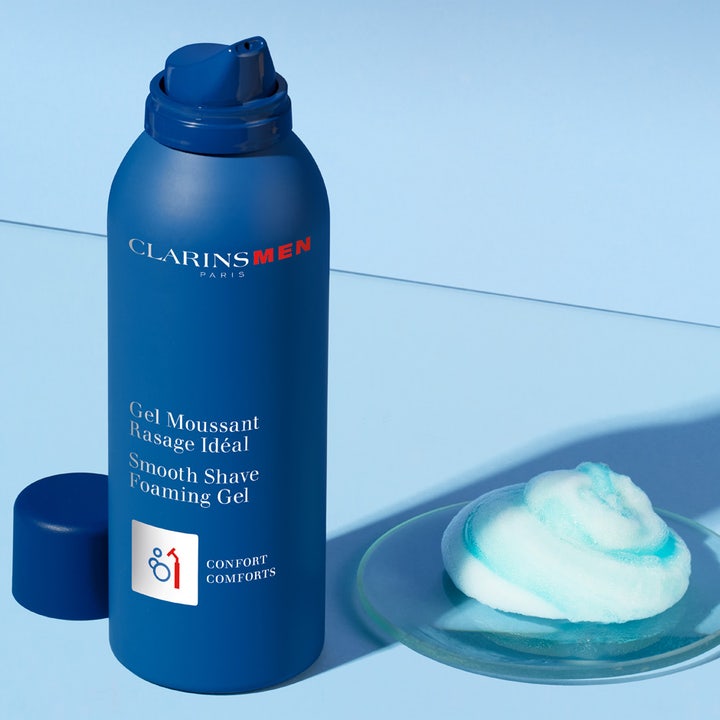 Clarins Clarins Men Shave Gel 150 ml rauhoittava parranajogeeli sinisessä pullossa ja ilmavaa vaahtoa lasialustalla.