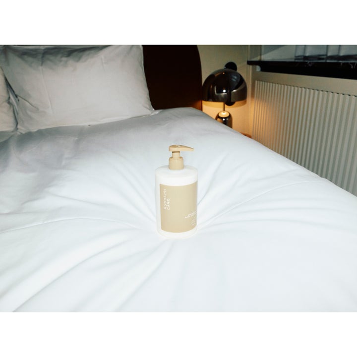 Rudolph Care Hand And Body Cream 500 ml pehmentävä vartalovoide pumppupullossa vaalealla vuoteella.