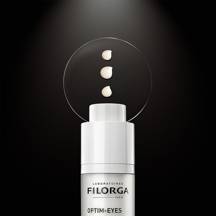 Filorga Optim-Eyes 3-in-1 15 ml silmänympärysvoide ja kolme voidepisaraa valkoisen pumppupullon yllä