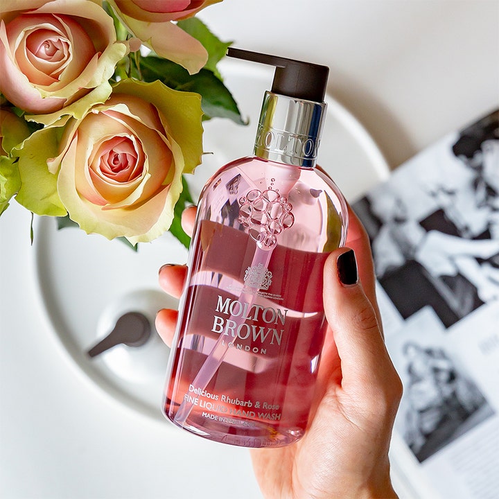 Molton Brown Delicious Rhubarb & Rose 300 ml puhdistava käsisaippua vaaleanpunaisessa pullossa ruusujen rinnalla.