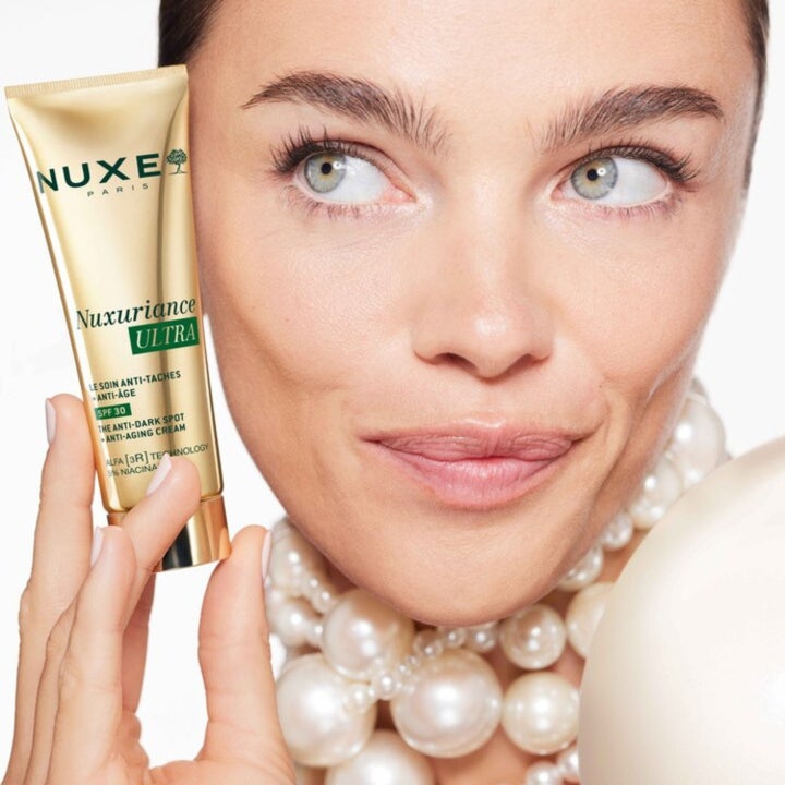 Nuxe Nuxuriance Ultra Anti Dark Spot Cream 50 ml päivävoide SPF 30 kultaisessa tuubissa helmiin pukeutuneen naisen kädessä.
