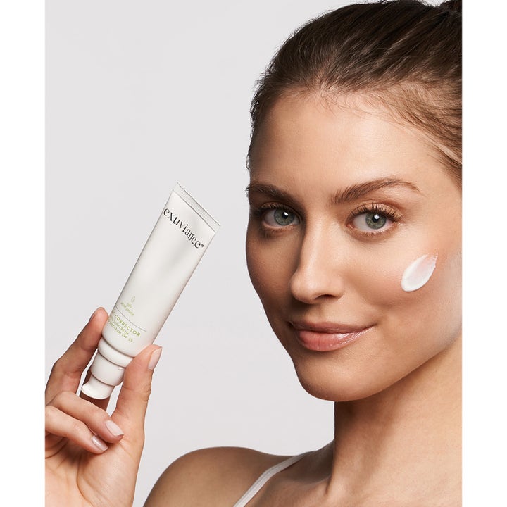Exuviance Daily Corrector SPF35 40 g tasoittava aurinkovoide valkoisessa tuubissa ja voidetta mallin poskella