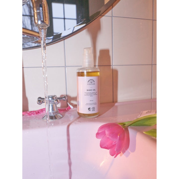 Rudolph Care Acai Body Oil 200 ml pehmentävä vartaloöljy pumppupullossa vaaleanpunaisen lavuaarin reunalla.