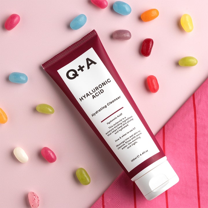 Q+A Hyaluronic Acid Gel Cleanser 125 ml kosteuttava puhdistusgeeli viininpunainen tuubi pinkillä taustalla karkkien kera