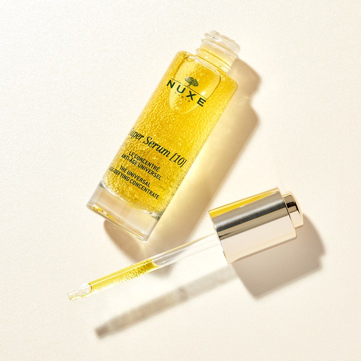 Nuxe Super Serum [10] 30 ml silottava anti-age-seerumi lasipullossa, jossa on kultaisia öljyhelmiä ja pipetti.