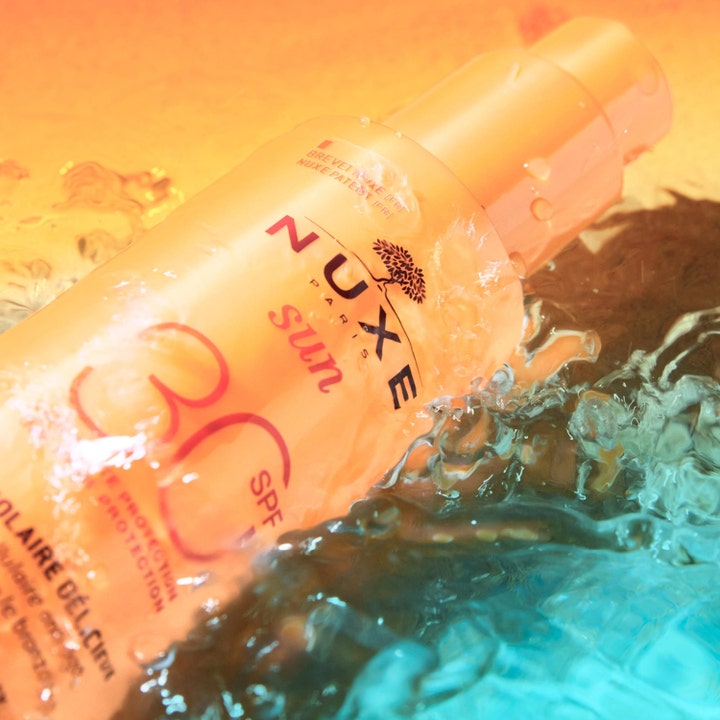 Nuxe Sun Spray SPF30 184 g aurinkosuoja kasvoille oranssissa pullossa veden loiskeiden ympäröimänä.