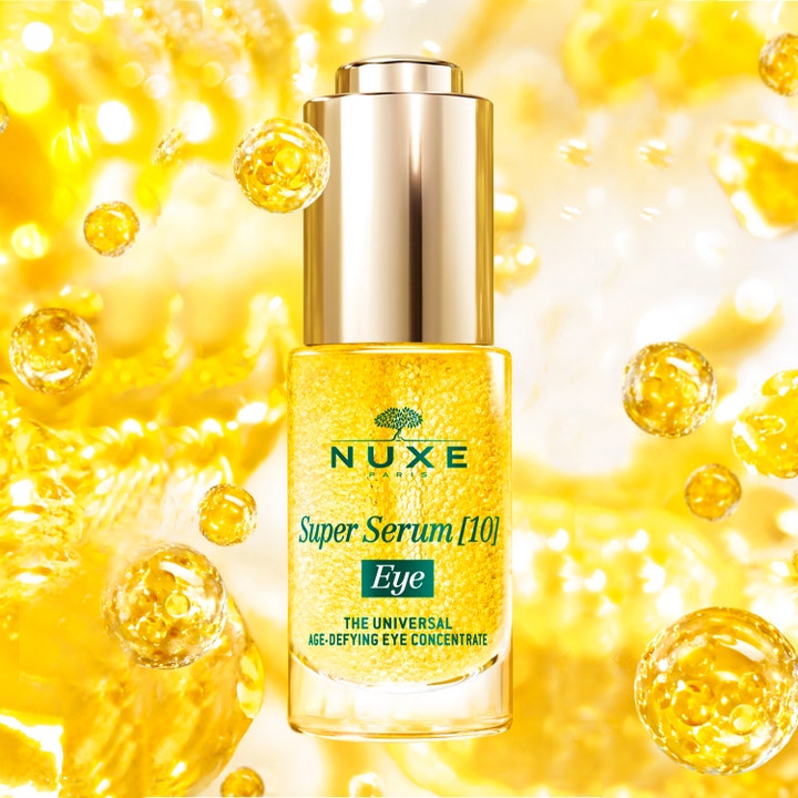 Nuxe Super Serum [10] Eye 15 ml silmänympärysseerumi kultaisessa pullossa keltaisten öljykuplien ympäröimänä.