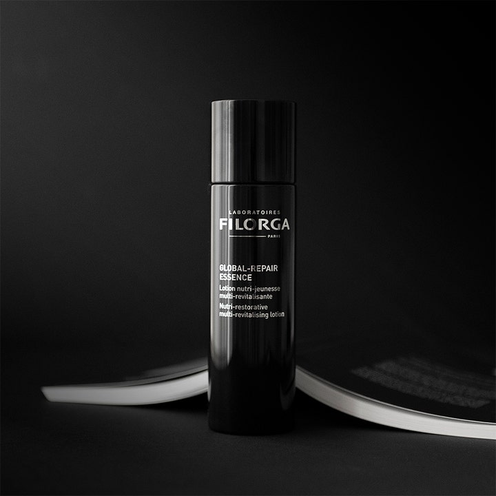 Filorga Global-Repair Essence 150 ml ravitseva hoitoneste mustassa lieriömäisessä pullossa avatun kirjan päällä