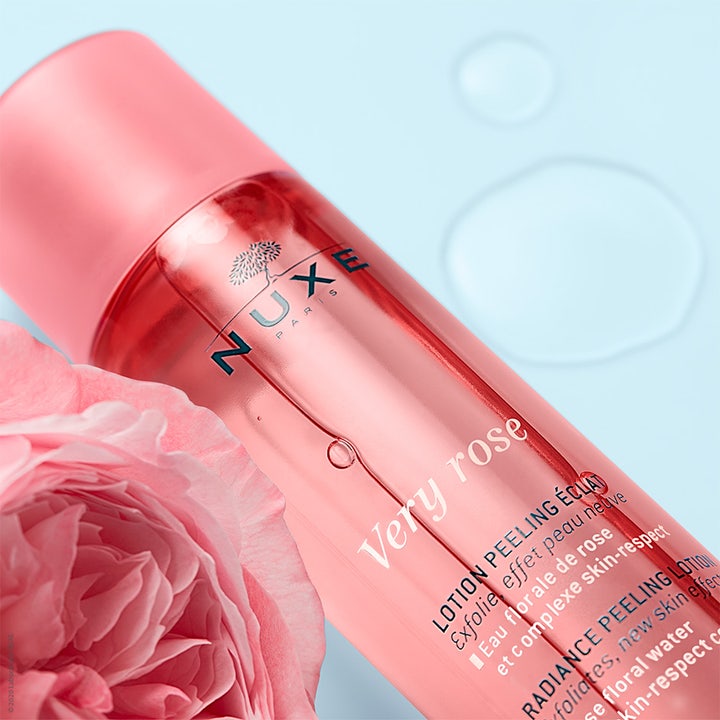 Nuxe Very Rose Peeling Lotion 150 ml heleyttävä kuorintavesi vaaleanpunaisessa pullossa ruusun ja veden äärellä.