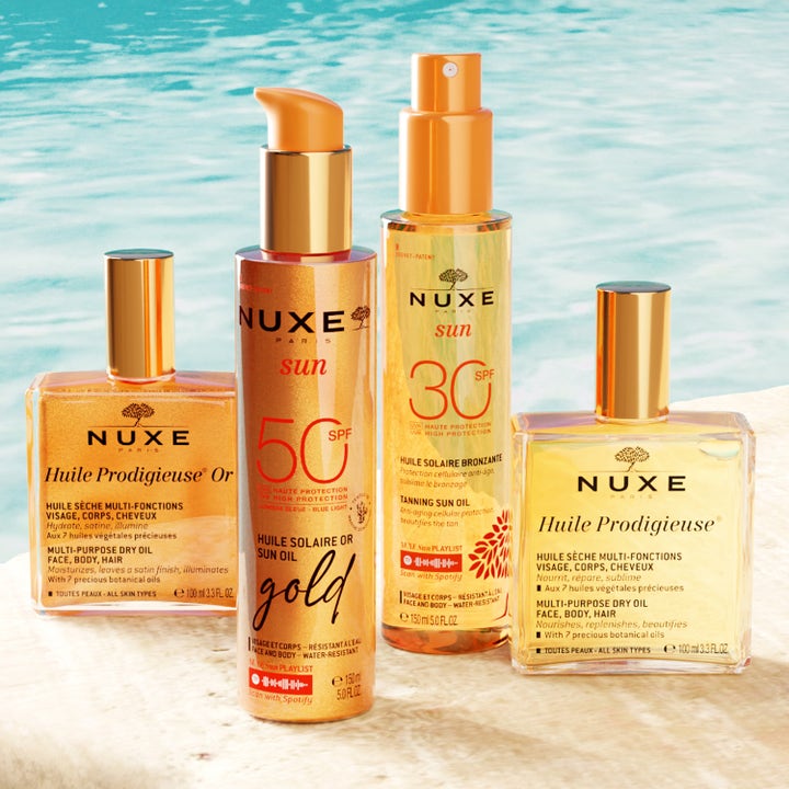 Nuxe Sun Tanning Oil Gold 150 ml hehkua antava aurinkosuojaöljy kultaisessa pumppupullossa veden äärellä.