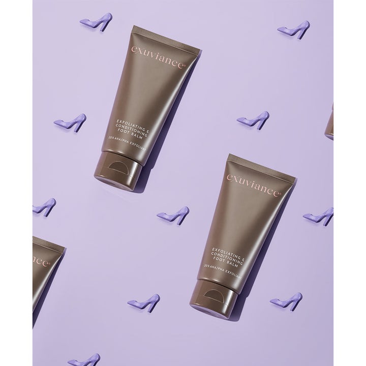 Exuviance Exfoliating & Conditioning Foot Balm 50 g kuoriva jalkavoide pronssituubeissa liilalla korkokenkätaustalla.