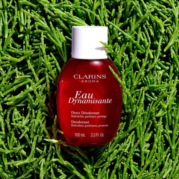 Clarins Eau Dynamisante Deodorant Spray 100 ml virkistävä deodorantti punaisessa pullossa vihreiden kasvien päällä.