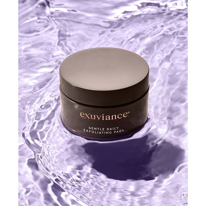 Exuviance Gentle Daily Exfoliating 60 Pads 55 ml kirkastava kuorinta mustassa purkissa liilassa vedessä.