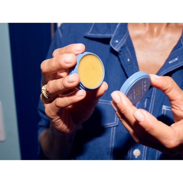 Rudolph Care Sun Balm SPF50 10 ml aurinkovoide on käsissä pidetty avattu sininen rasia, jossa näkyy keltainen voide