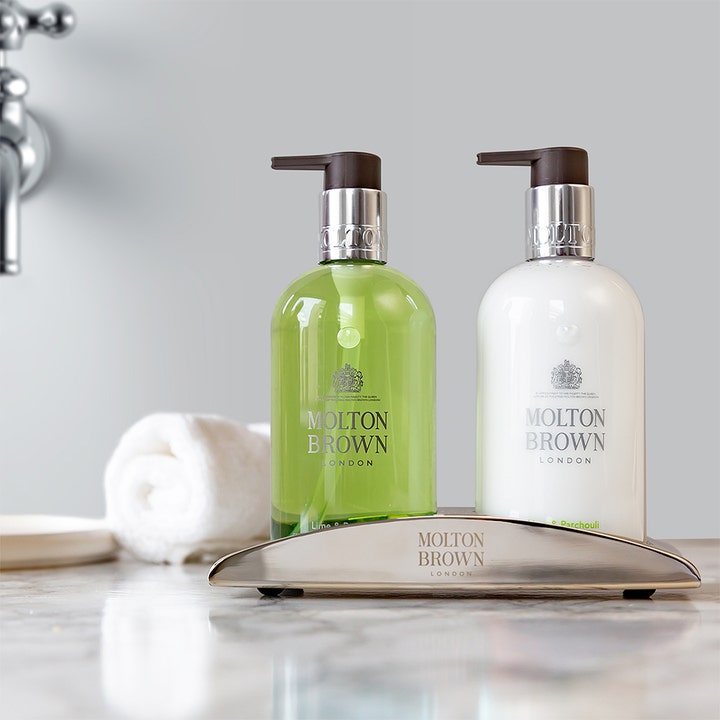 Kosteuttava Molton Brown Lime & Patchouli Hand Lotion 300 ml käsivoide valkoisessa pumppupullossa telineessä.