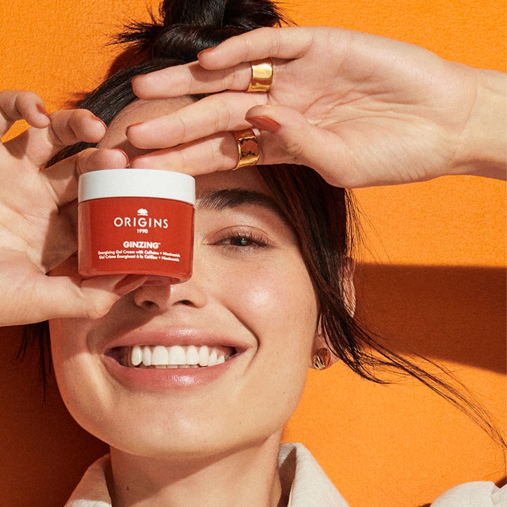 Origins Ginzing Energizing Gel Face Cream 50 ml kirkastava kasvovoide jota hymyilevä nainen pitelee oranssia vasten.