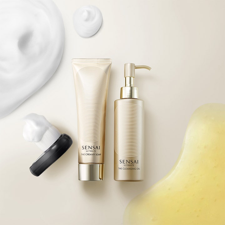 Sensai Ultimate The Cleansing Oil 150 ml puhdistusöljy kultaisessa pumppupullossa vaaleiden tekstuurien rinnalla