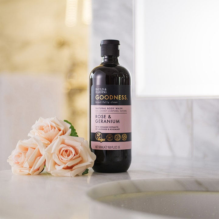 Baylis & Harding Goodness Rose & Geranium 500 ml suihkugeeli tummassa pullossa vaaleanpunaisten ruusujen vieressä.